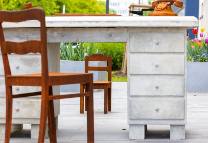ein Schreibtisch aus Beton mit einem großen Stuhl, auf der einen und einem kleinen Kinderstuhl auf der gegenüberliegenden Seite