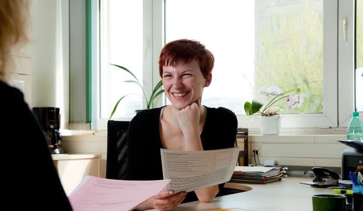 Foto: zwei Frauen sitzen sich an einem Schreibtisch in einem Beratungsgespräch gegenüber und haben Zettel in der Hand