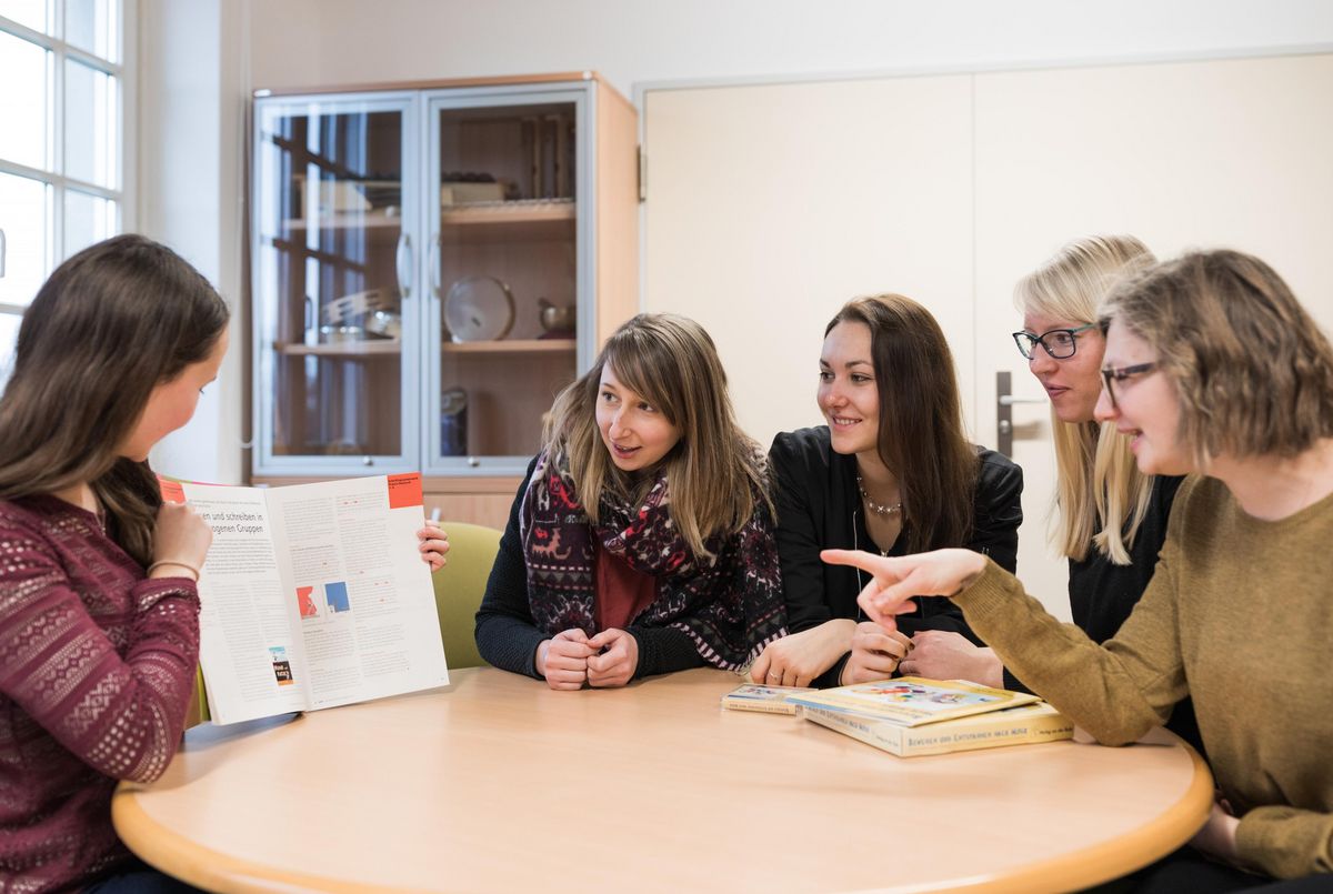 zur Vergrößerungsansicht des Bildes: Fünf Studierende sitzen an einem runden Tisch. Links hält eine Studierende ein aufgeschlagenes Buch in der Hand und zeigt es den anderen Studierenden.