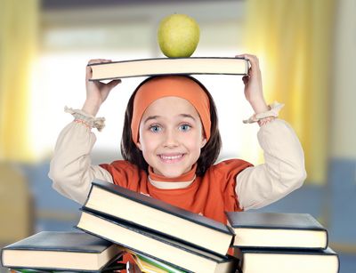 Ein Mädchen lächelt in die Kamera und hält ein Buch über dem Kopf, auf dem ein Apfel liegt.