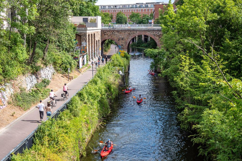zur Vergrößerungsansicht des Bildes: Der Karl-Heine-Kanal mit einer der zahlreichen Brücken in Leipzig. Im Wasser paddeln mehrere Kanus, auf dem Fußweg daneben spazieren Fußgänger.