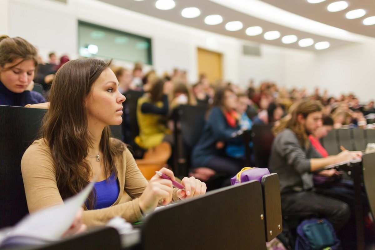 zur Vergrößerungsansicht des Bildes: Fokus auf eine Studentin in der Vorlesung. Unscharf im Hintergrund sieht man den vollen Hörsaal, Foto: Christian Hüller 