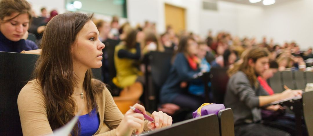 Fokus auf eine Studentin in der Vorlesung. Unscharf im Hintergrund sieht man den vollen Hörsaal, Foto: Christian Hüller 