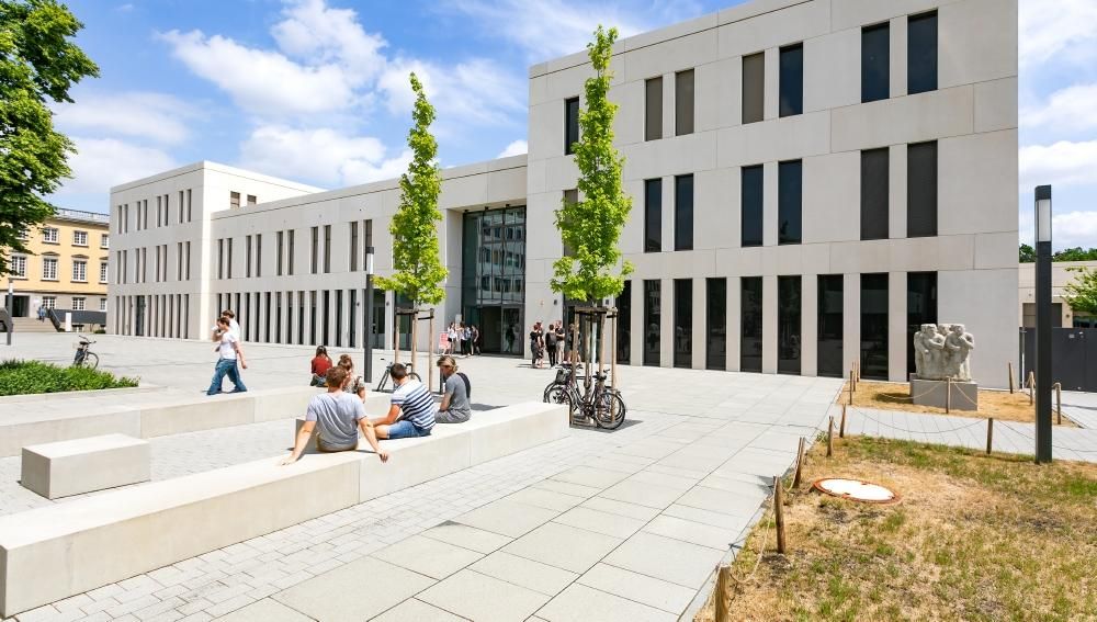 Ein modernes, beigefarbenes Gebäude, davor sitzen auf dem Campus Studierende.