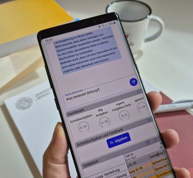 Hand hält ein Smartphone, auf dem der Chatbot zu sehen ist. Die eingetippte Frage lautet: "Was bedeutet Bildung?"
