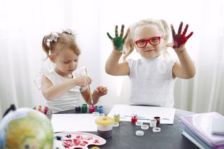 Zwei Kinder spielen mit Farbtöpfen. Eines der Kinder hält ihre bunten Finger in die Kamera.