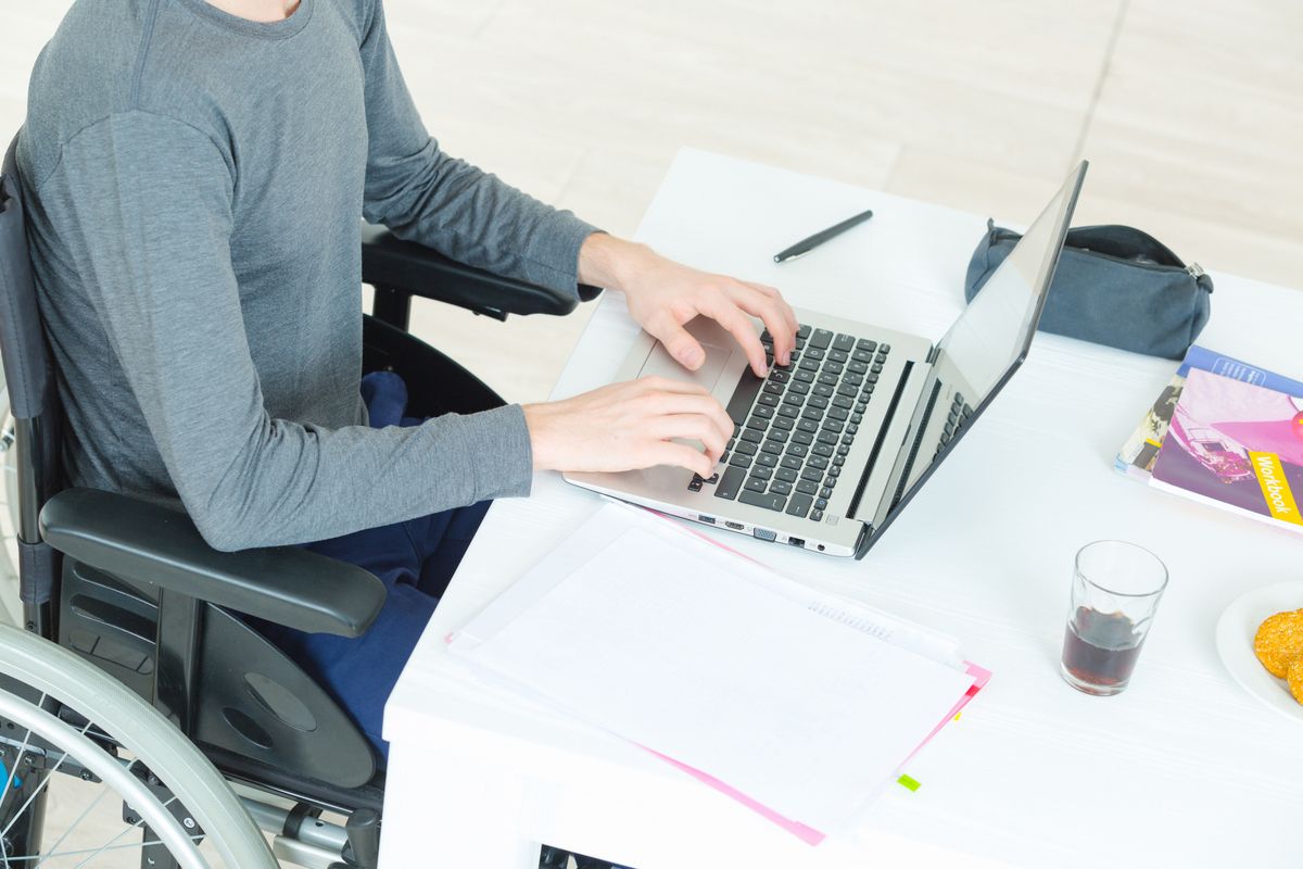 zur Vergrößerungsansicht des Bildes: Ein Mensch im Rollstuhl arbeitet an einem Schreibtisch am Laptop.