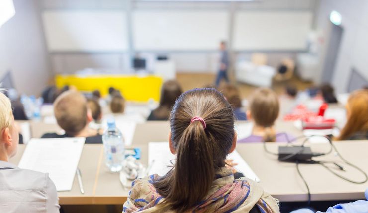 Ein Vorlesungssaal. Im Vordergrund die Hinterköpfe der Studierenden, im Hintergrund verschwommen der Vortragende am anderen Ende des Raumes.