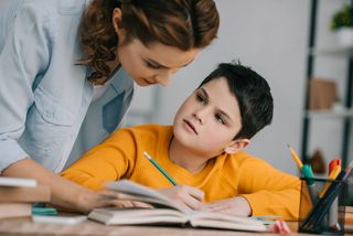 Junge bearbeitet seine Hausaufgaben zu Hause. Seine Mutter schaut ihm über die Schultern, um ihm zu helfen.