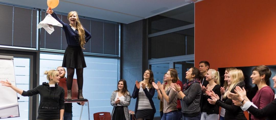 Seminarraum: Eine Person steht auf einen Stuhl, der auf einen Tisch steht und hält einen orangenen Luftballon in die Höhe. Sie blickt freudig auf ihn. Rechts im Bild steht eine Gruppe Studierender, die zu ihr blicken und applaudieren, Foto: Christian Hüller