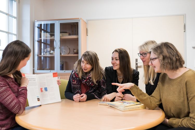 Fünf Studierende sitzen um einen runden Tisch und schauen sich ein Lehrbuch an, welches von einer Studentin, die mit am Tisch sitzt, hochgehalten wird für alle, Foto: Christian Hüller