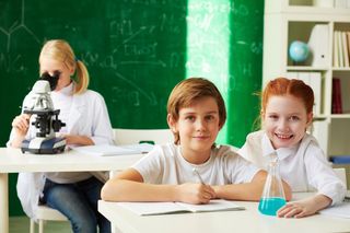 Auf dem Bild sind drei Kinder abgebildet. Zwei von ihnen sitzen zusammen an einem Tisch und schreiben etwas in ihr Heft. Auf ihrem Tisch steht außerdem ein Kolben mit blauer Flüssigkeit. Hinter dem Tisch der beiden sitzt ein weiteres Kind an einem anderen Tisch. Es schaut in ein Mikroskop. Hinter den Kindern befindet sich eine Tafel, die mit Formeln beschrieben ist, sowie ein Regal mit vielen Unterlagen und einem Globus.