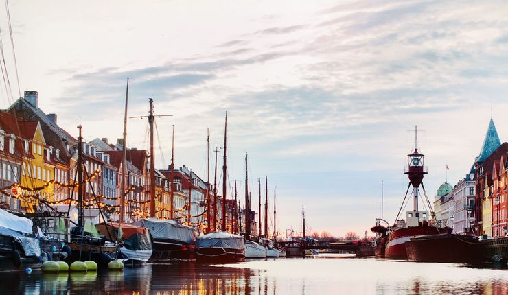 Der Hafen Nyhavn in Kopenhagen, die bunten Häuser und Boote spiegeln sich im Wasser. Lichterketten leuchten in der beginnenden Dämmerung.