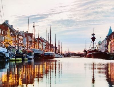 Der Hafen Nyhavn in Kopenhagen, die bunten Häuser und Boote spiegeln sich im Wasser. Lichterketten leuchten in der beginnenden Dämmerung.