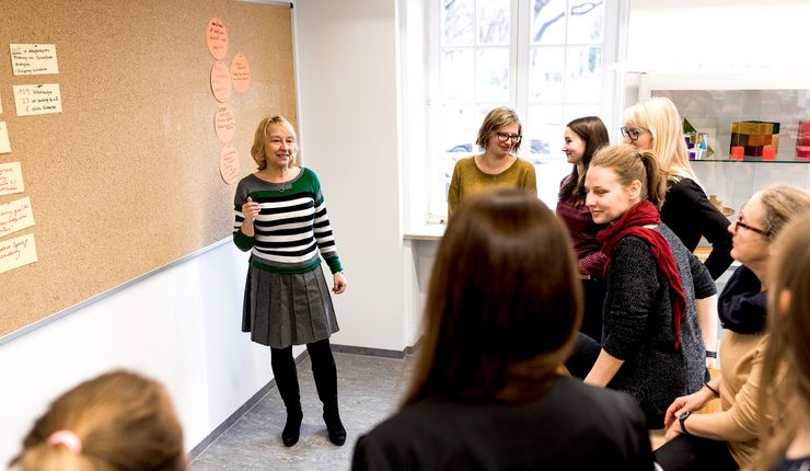 Professor Doktor Susanne Viernickel steht vor einer großen Pinnwand mit Moderationskarten in einem Seminarraum. Studierende stehen vor der Pinnwand, schauen in Richtung der Professorin und sprechen zum Teil miteinander.