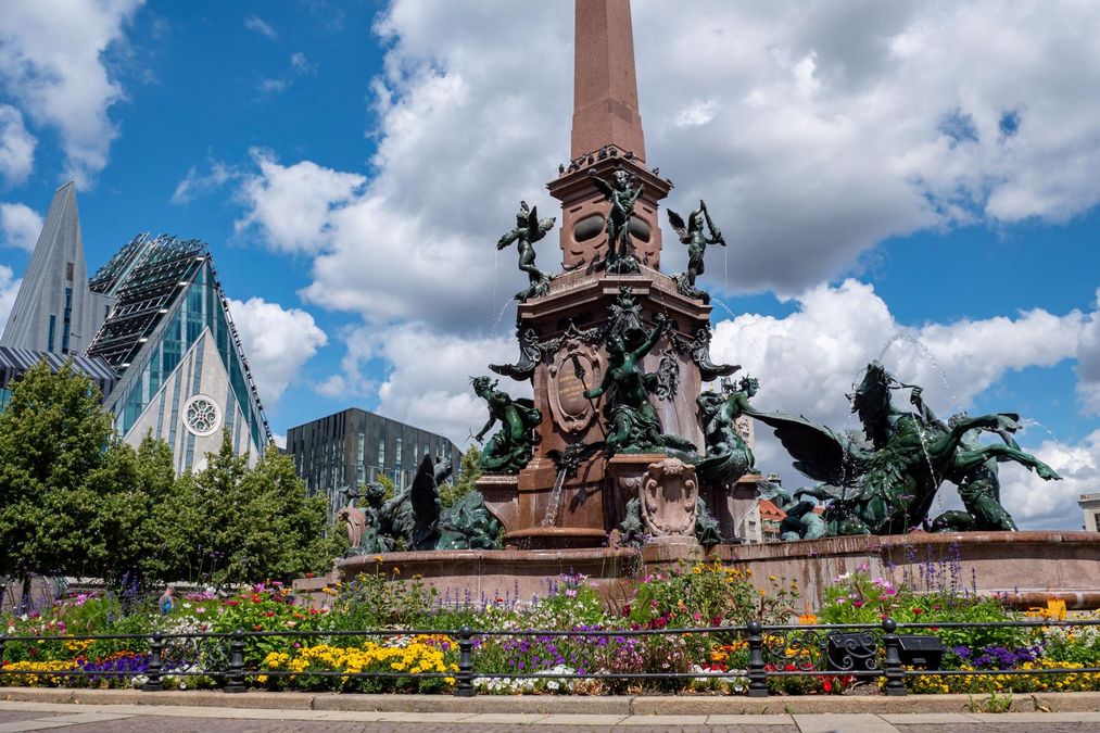 zur Vergrößerungsansicht des Bildes: Ein mit großen Steinfiguren verzierter Brunnen, im Hintergrund das futuristische Kirchengebäude der Uni Leipzig.