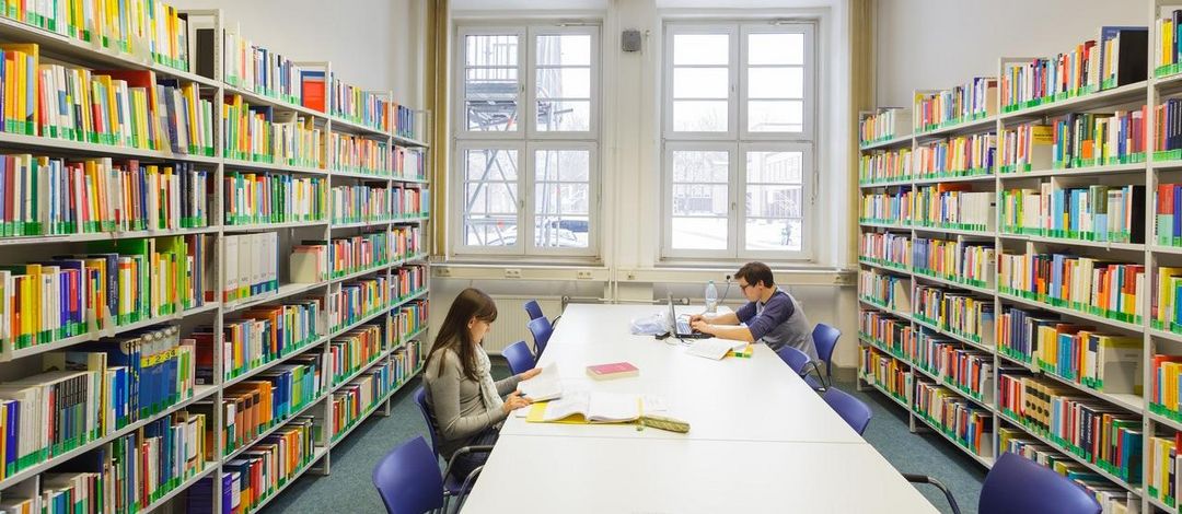 Blick auf zwei Studierende, die an einem langen Tisch zwischen großen Bücherregal sitzen, Foto: Christian Hüller 