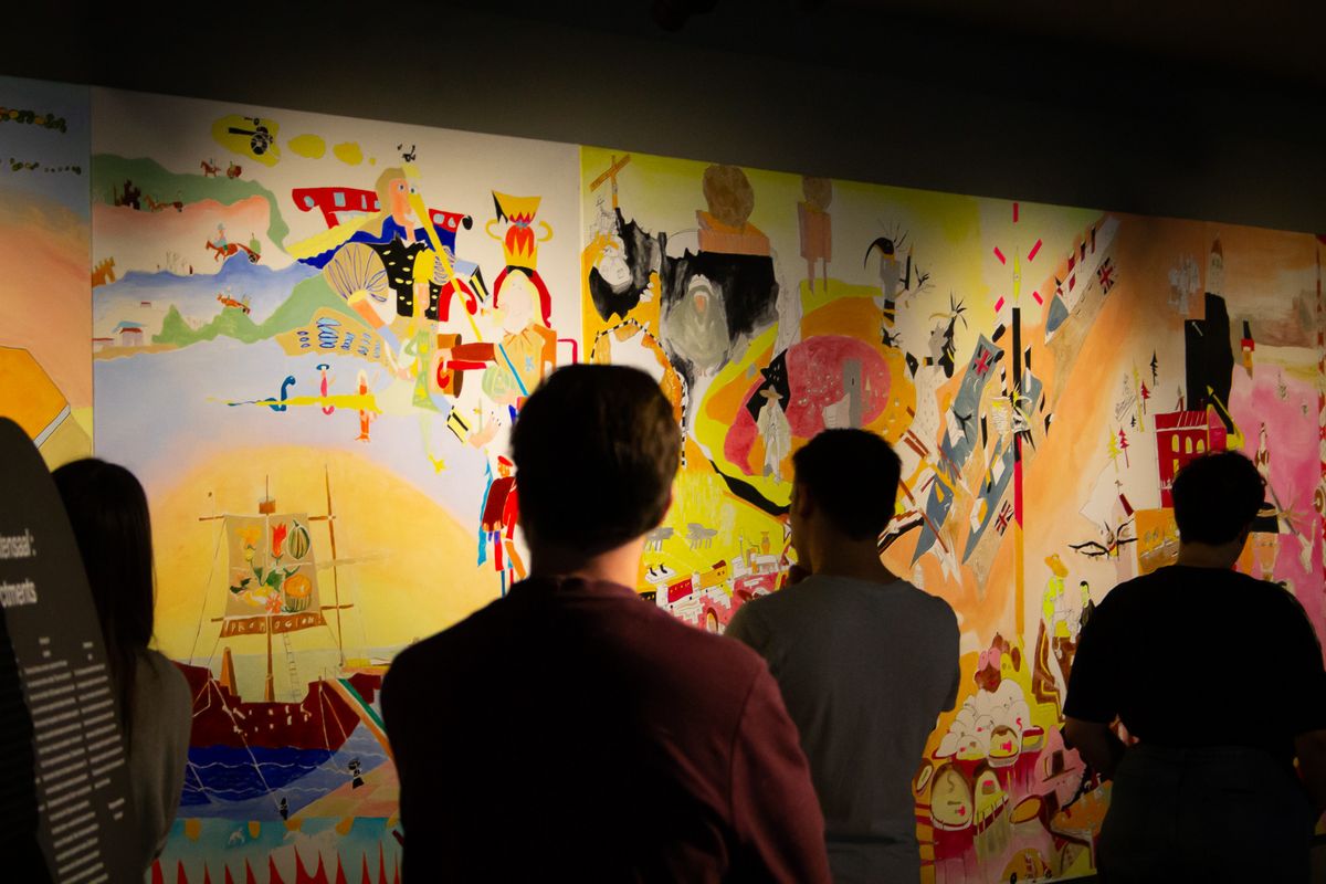 zur Vergrößerungsansicht des Bildes: Das Bild zeigt eine Gruppe von Studierenden in einer Ausstellung. Sie betrachten die im Hintergrund aufgetragene Graphic Novel.