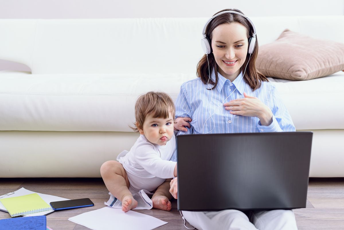 zur Vergrößerungsansicht des Bildes: Eine Frau sitzt mit Headset und Laptop auf dem Schoß auf dem Boden, während ein Kleinkind neben ihr sitzt.