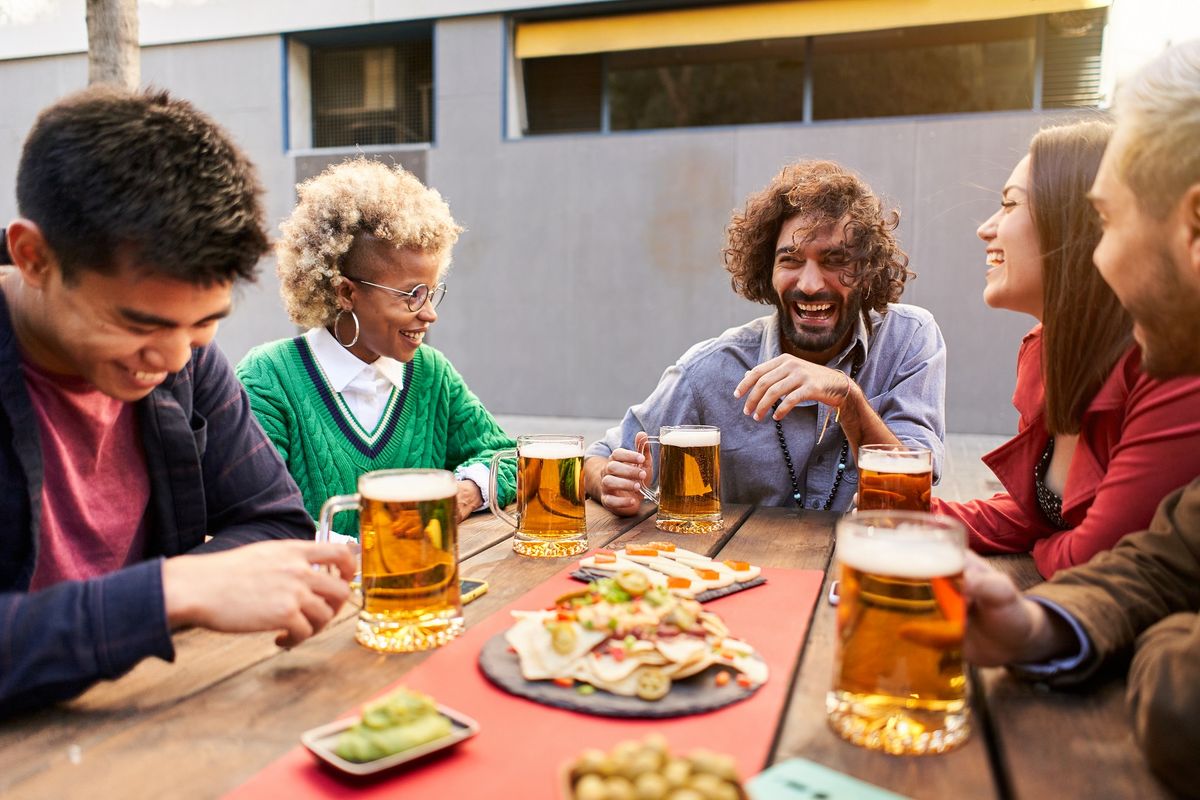zur Vergrößerungsansicht des Bildes: Auf dem Bild sind Menschen sitzend an einem Tisch zu sehen. Sie essen, trinken und lachen.