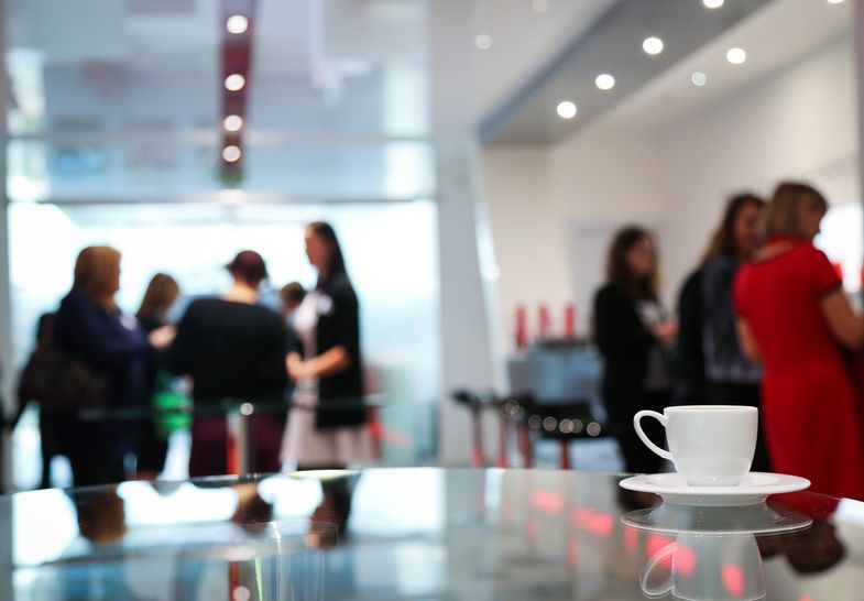 Eine Kaffeepause während einer Konferenz: Im Vordergrund ist eine Kaffeetasse auf einem Glastisch zu sehen. Im Hintergrund unscharf sind zwei Gruppen von Menschen zu sehen, die sich unterhalten. 