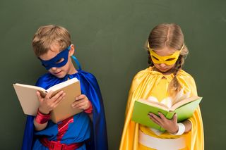 2 Kinder im Superheldenkostüm vor einer Tafel stehend mit Büchern in der Hand