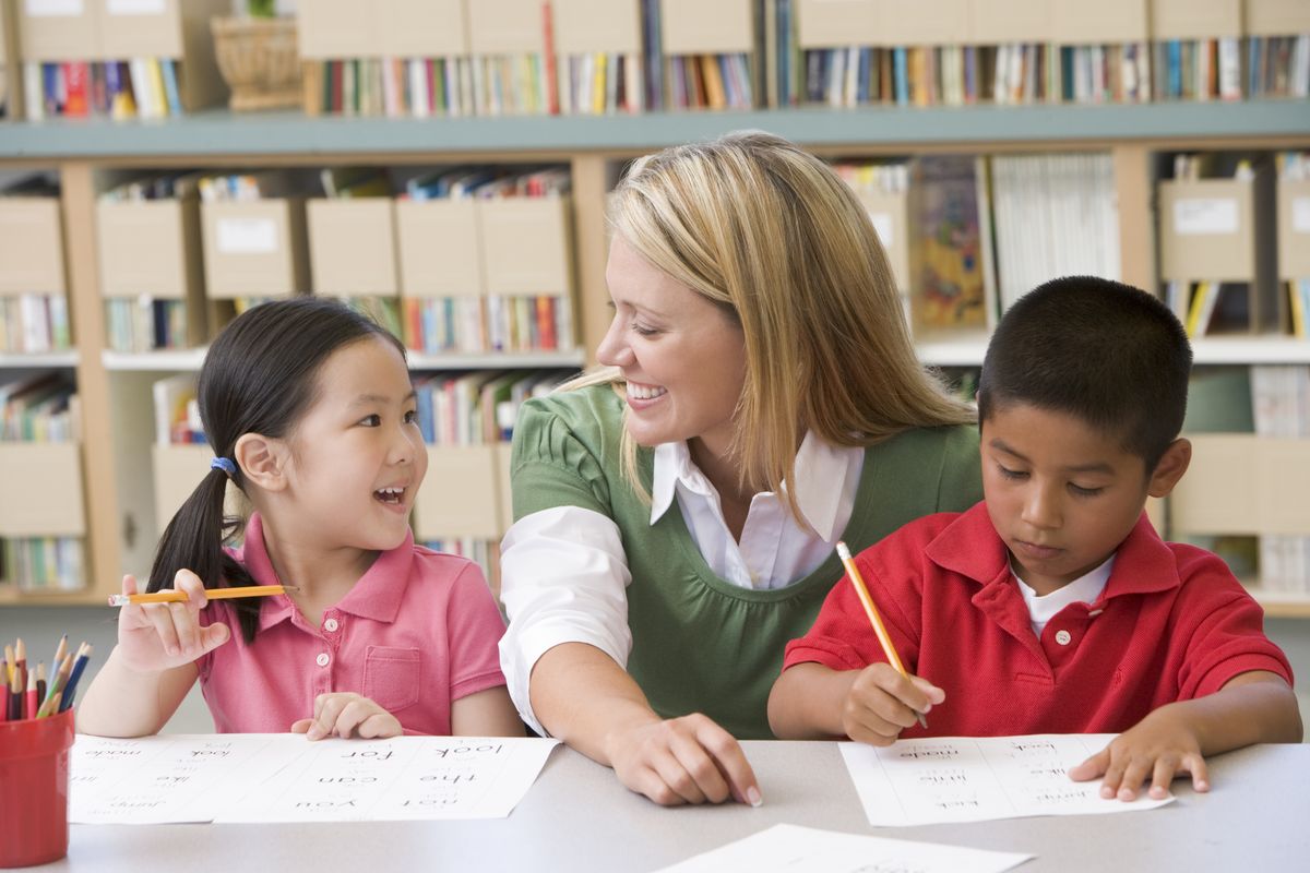 enlarge the image: Eine Lehrerin hockt an einem Tisch mit zwei Kindern und hilft ihnen beim Lernen.