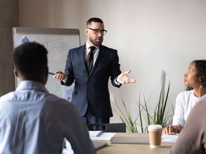 Eine Gruppe Seminarteilnehmender hört einem Vortrag zu. Der Redner steht an einem Flipchart und gestikuliert.