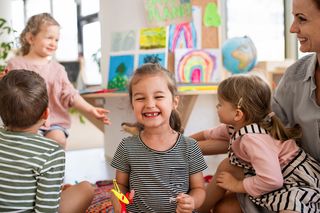 eine Fachkraft sitzt mit vier Kindern auf dem Boden, ein Kind iin der Mitte grinst in die Kamera