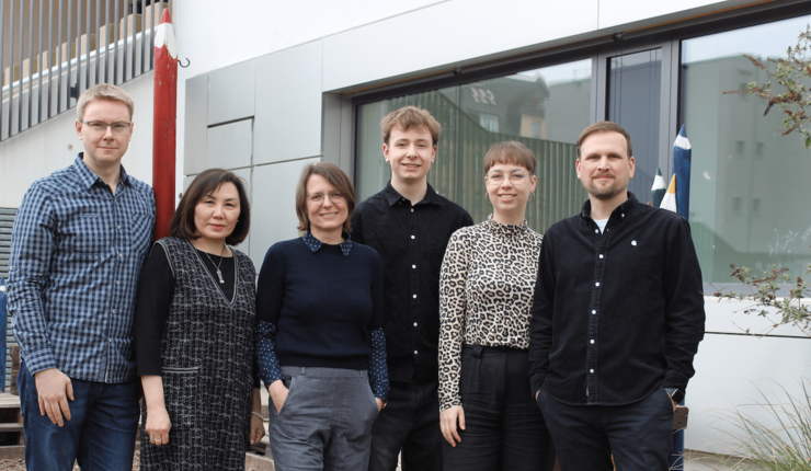 Farbfoto mit 6 Personen, davon drei Männer und drei Frauen vor einer Gebäudewand, alle Personen schauen in die Kamera und lächeln